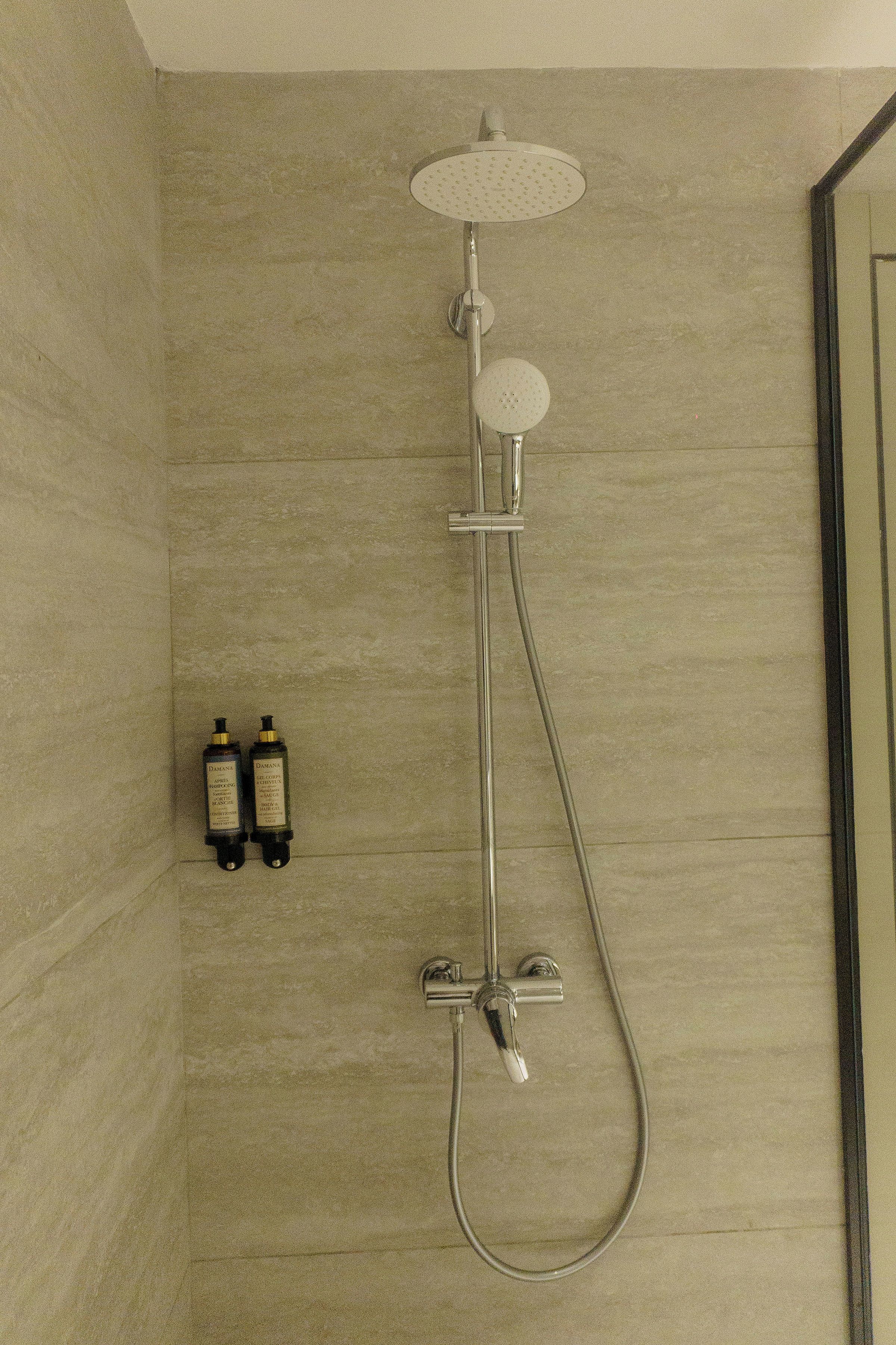 A shower with a handheld showerhead and a wall-mounted faucet. There are two bottles of shampoo and conditioner on the wall.