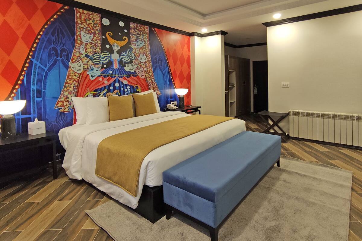 A hotel room with a large bed, a bench, a nightstand with a lamp, a wall tapestry, and a radiator.