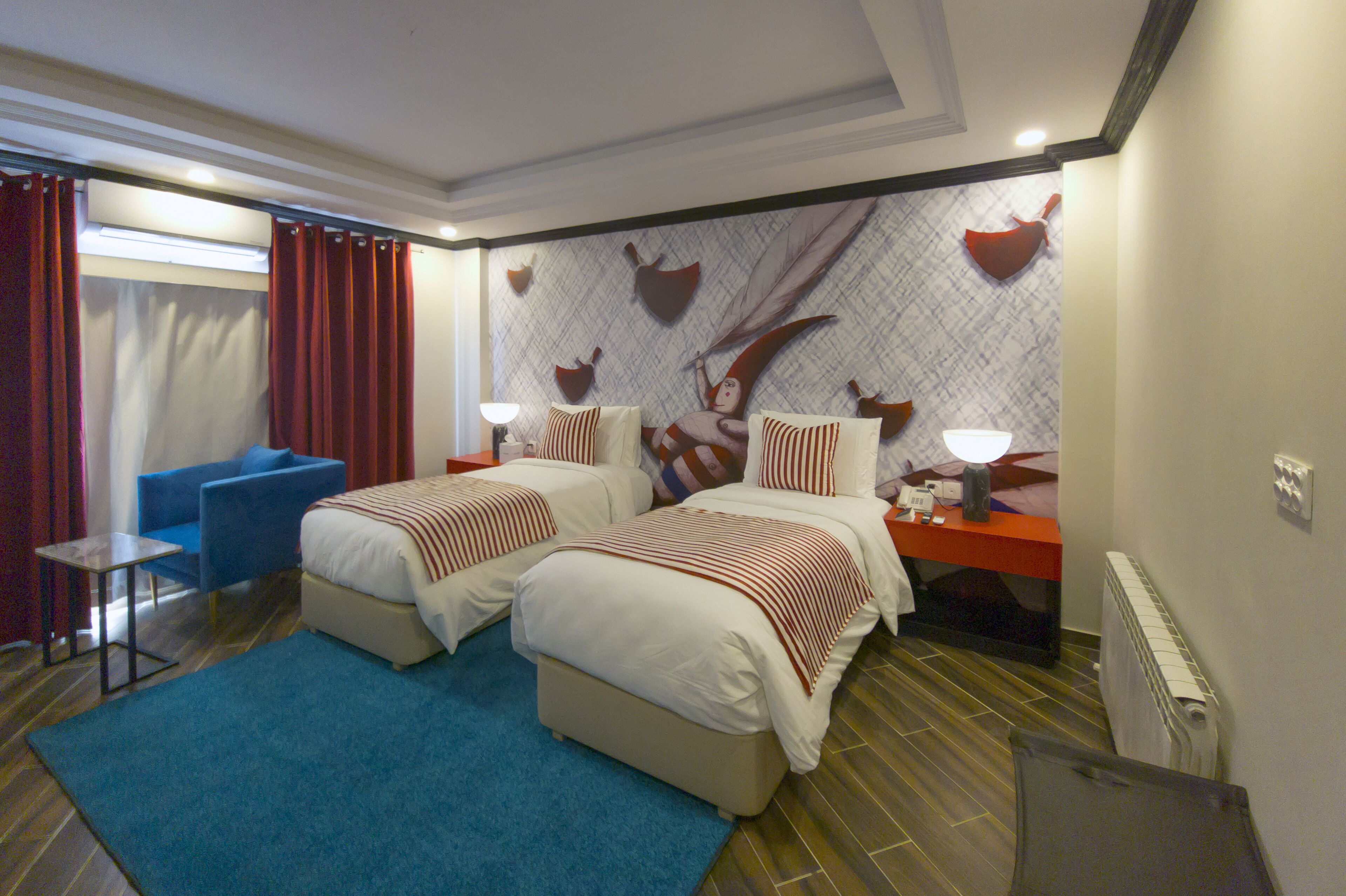 A hotel room with two beds, a blue armchair, a small table, and a wall with decorative elements.