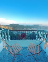 Un balcón con dos sillas de madera y baranda de metal decorativa, con vistas a un paisaje montañoso.