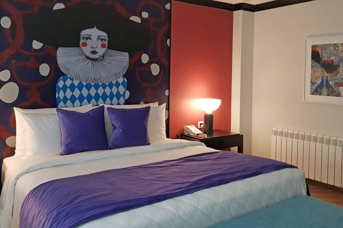 A hotel room with a bed, a nightstand, a lamp, a radiator, and a large abstract painting on the wall.