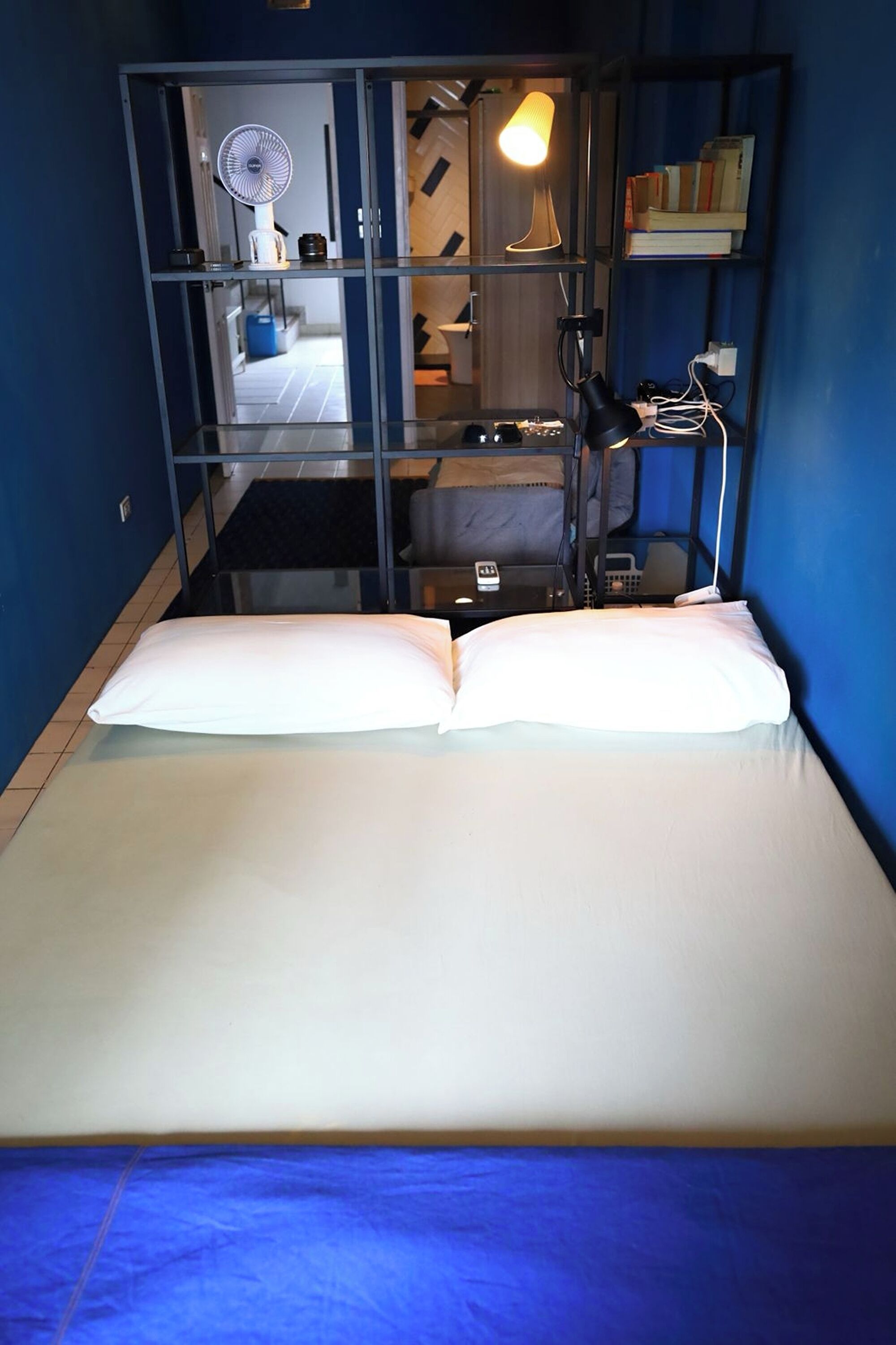 Una cama con almohadas blancas y una colcha azul, un escritorio con una lámpara, una estantería y un espejo que refleja una habitación con un ventilador y una lámpara.