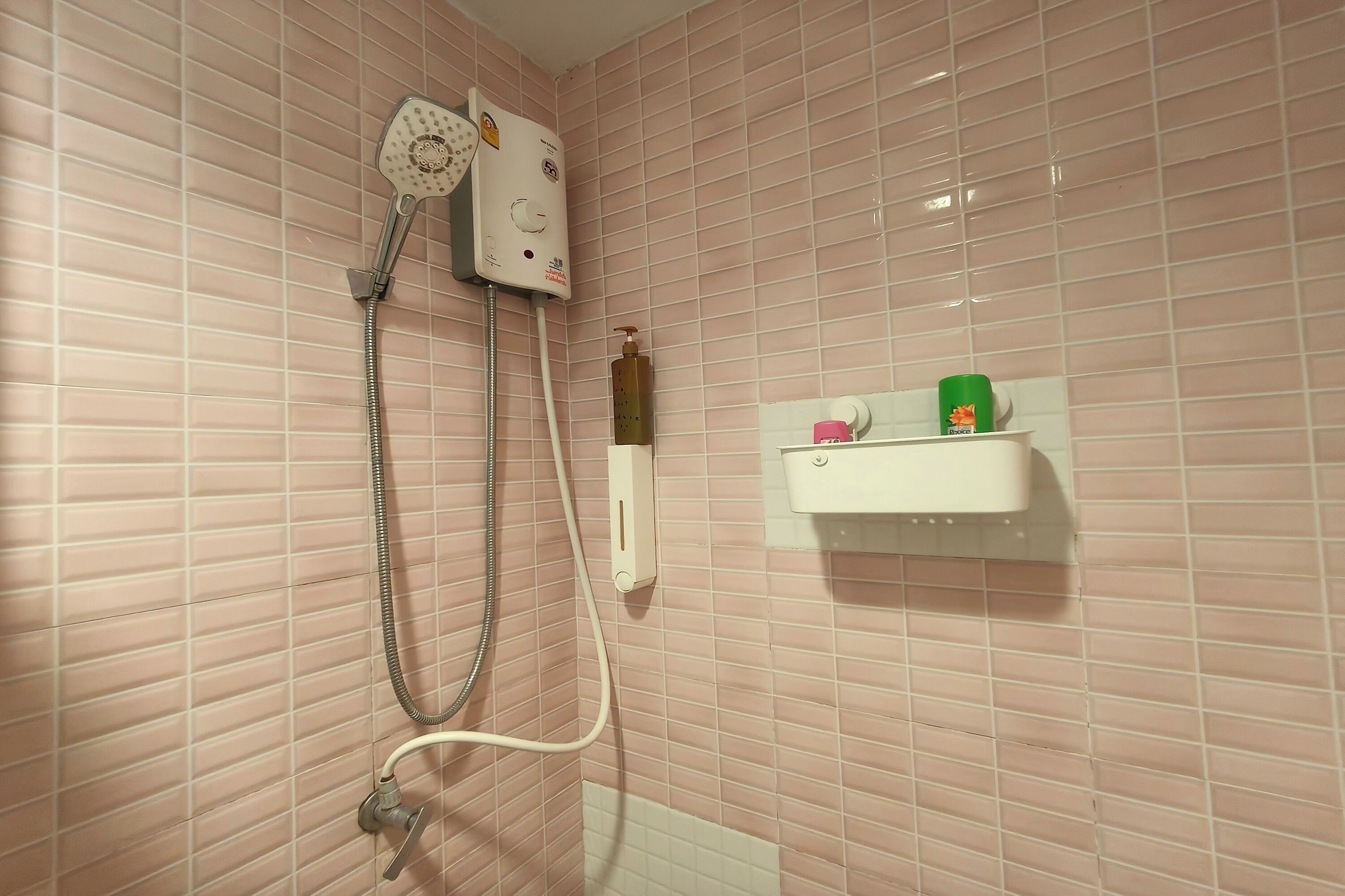 A bathroom with pink tiled walls, a shower with a handheld showerhead, and a small shelf with toiletries.