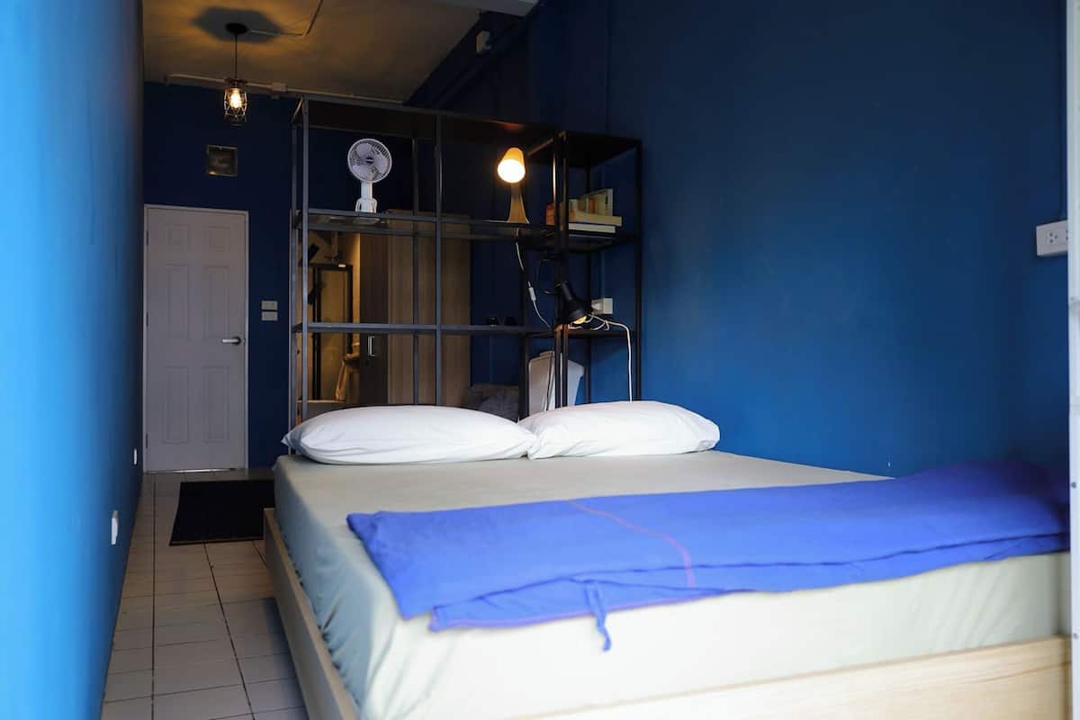 A bedroom with a bed, a bedside table, a lamp, and a shelf with books and a fan.