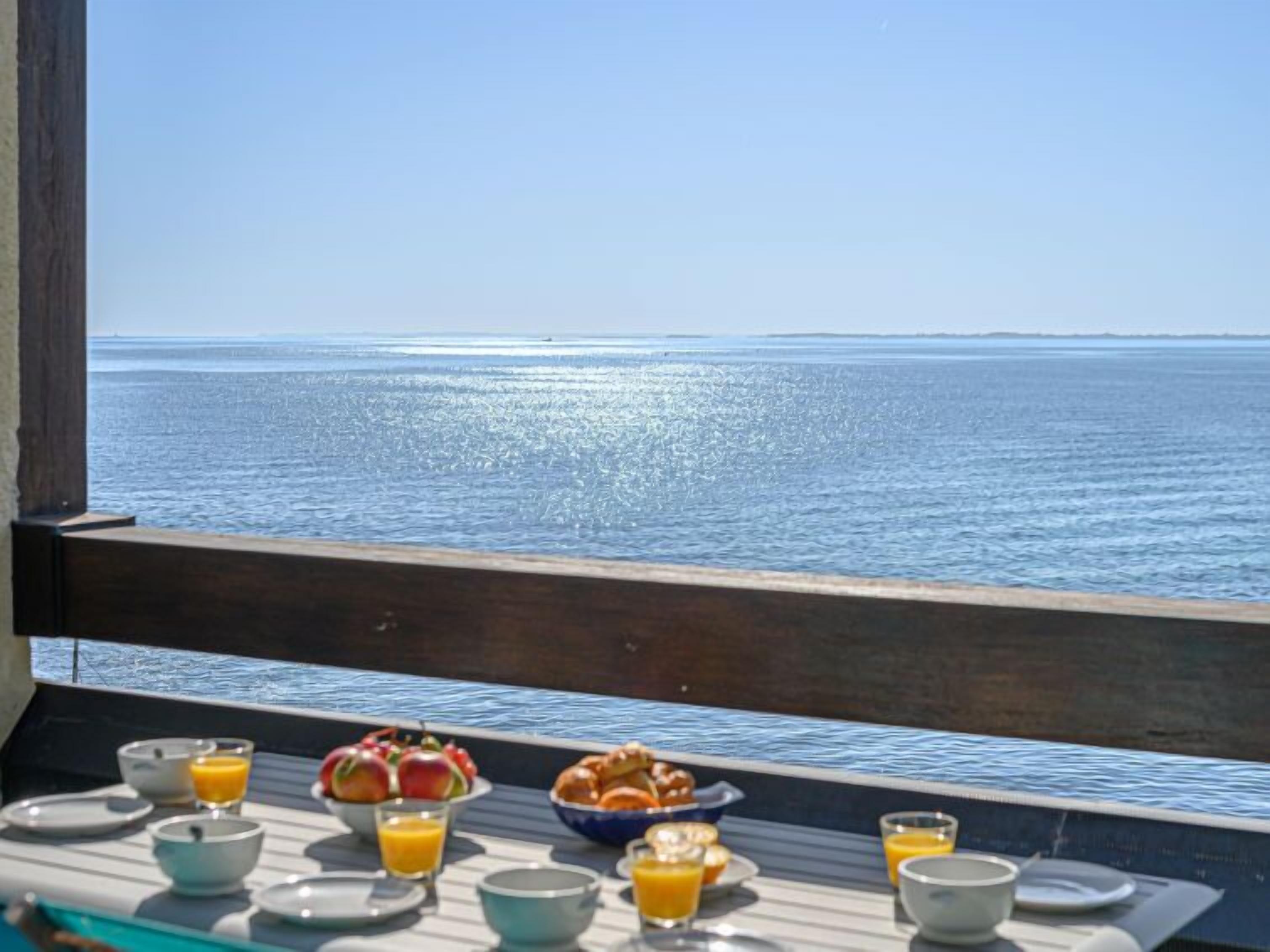 Une table de petit-déjeuner avec des fruits, des viennoiseries et du jus d’orange, donnant sur une mer calme.
