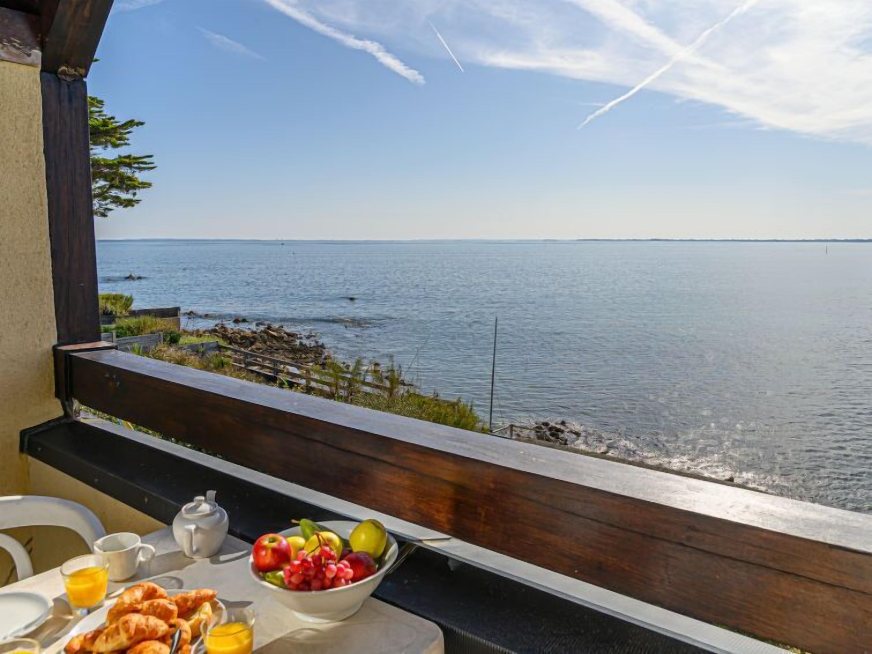 Une table de petit-déjeuner avec des croissants, des fruits et du jus d’orange, placée sur un balcon donnant sur une mer calme.