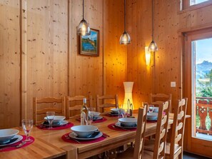 A wooden dining room with a table set for six, two pendant lights, a painting on the wall, and a view of mountains through a window.