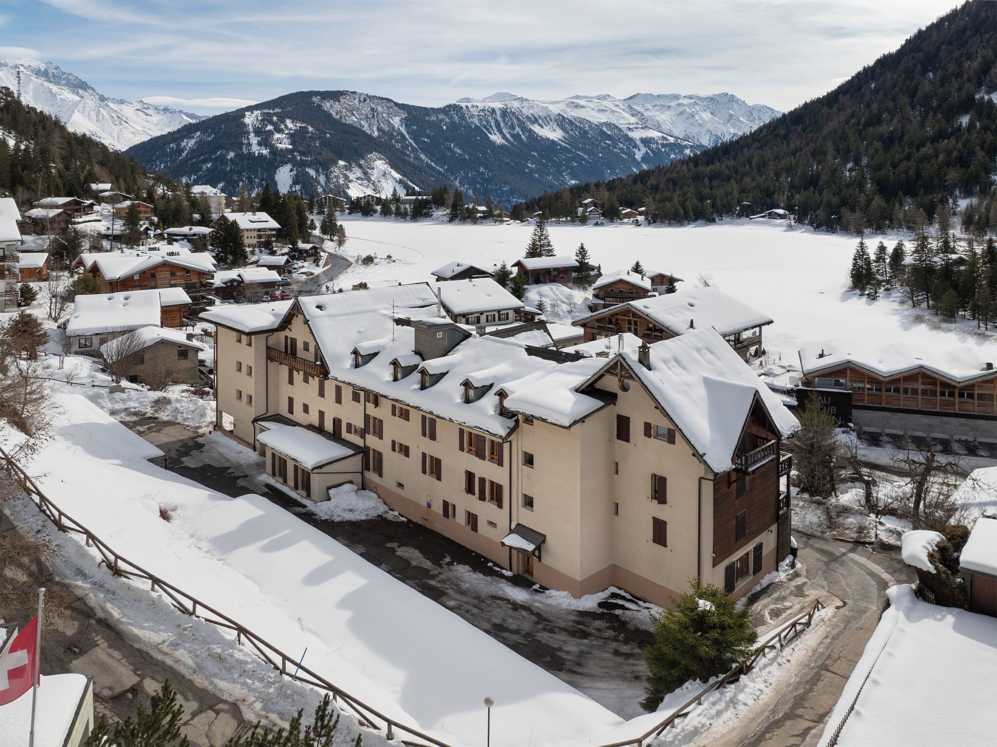 A snow-covered mountain town with a large hotel featuring multiple buildings, a ski lift, and a view of snow-capped mountains.
