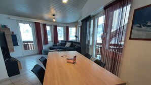 Ein geräumiges Zimmer mit einem großen Holztisch, schwarzen Stühlen, einer Couch und einem Balkon mit roten Vorhängen.
