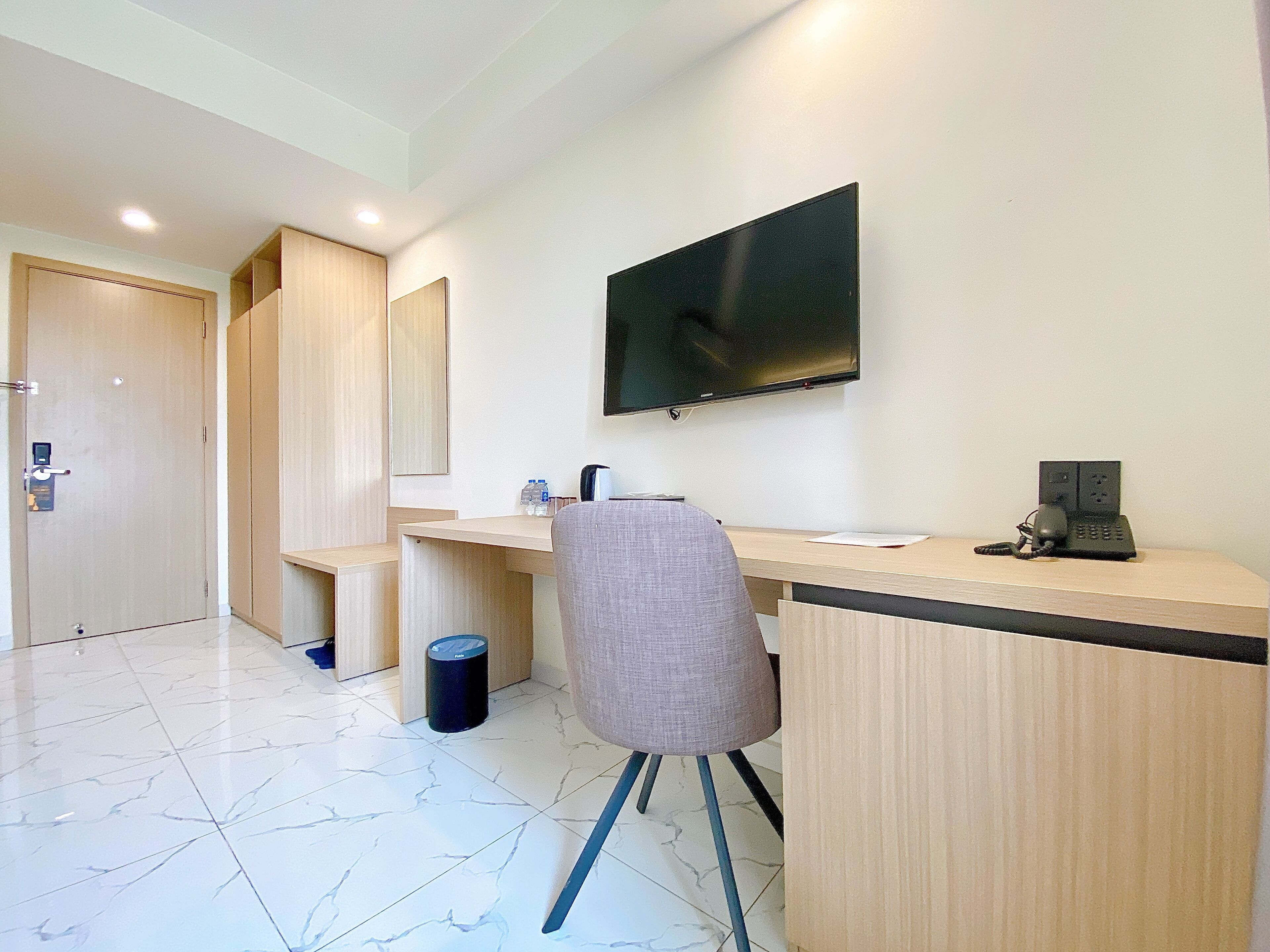 A modern hotel room with a wooden desk, a chair, a flat-screen TV mounted on the wall, and a trash bin.