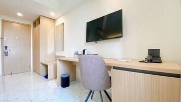 A modern hotel room with a wooden desk, a chair, a flat-screen TV mounted on the wall, and a trash bin.