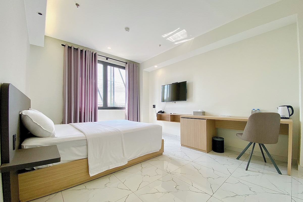 Una habitación de hotel con cama, escritorio, silla, televisor y ventana con cortinas.