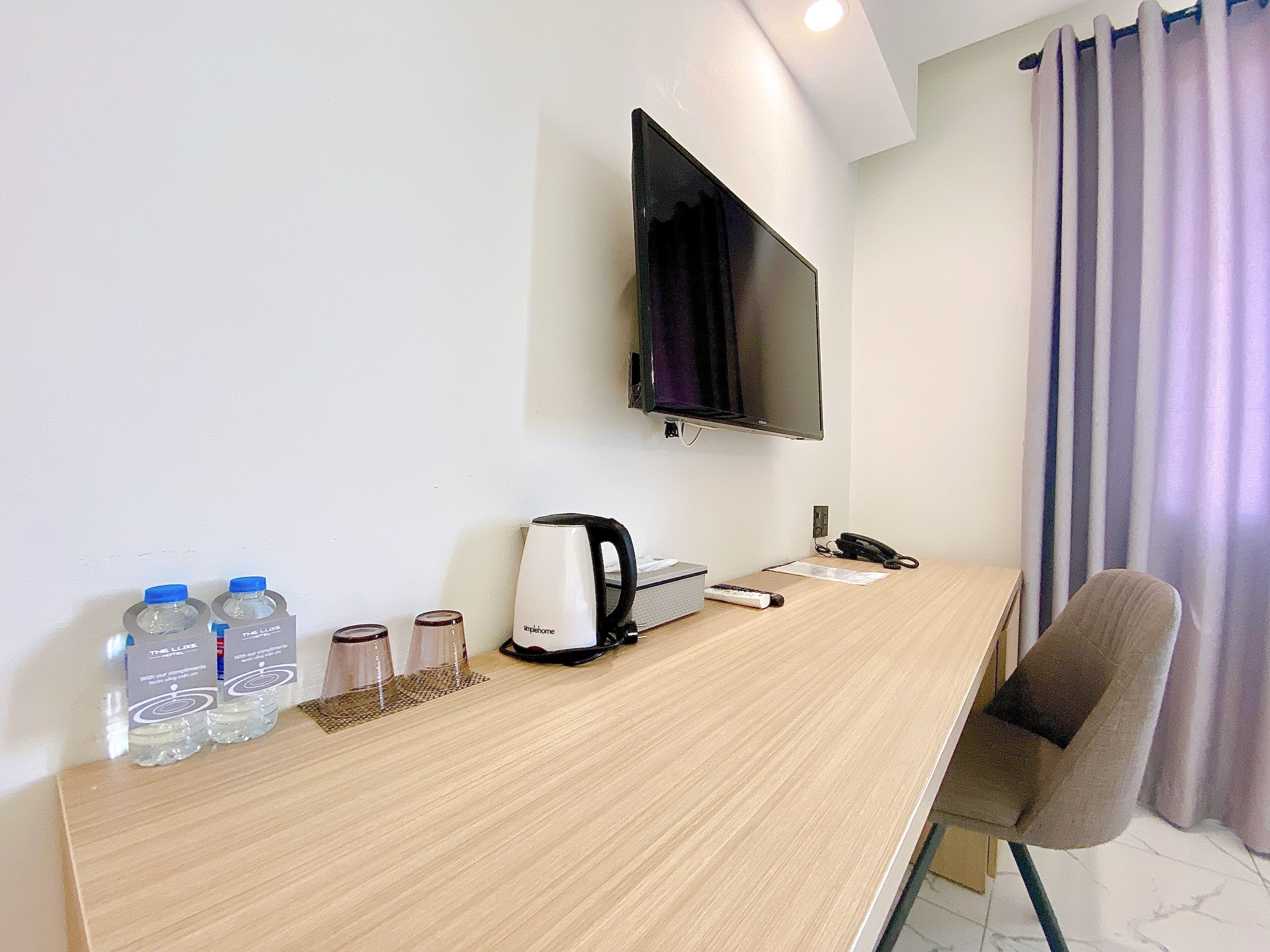 Quarto de hotel com escrivaninha de madeira, televisão de tela plana na parede, cadeira, controle remoto, chaleira e água engarrafada.