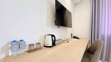 Quarto de hotel com escrivaninha de madeira, televisão de tela plana na parede, cadeira, controle remoto, chaleira e água engarrafada.