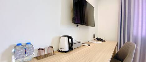 Habitación de hotel con un escritorio de madera, un televisor de pantalla plana montado en la pared, una silla, un control remoto, una tetera y agua embotellada.