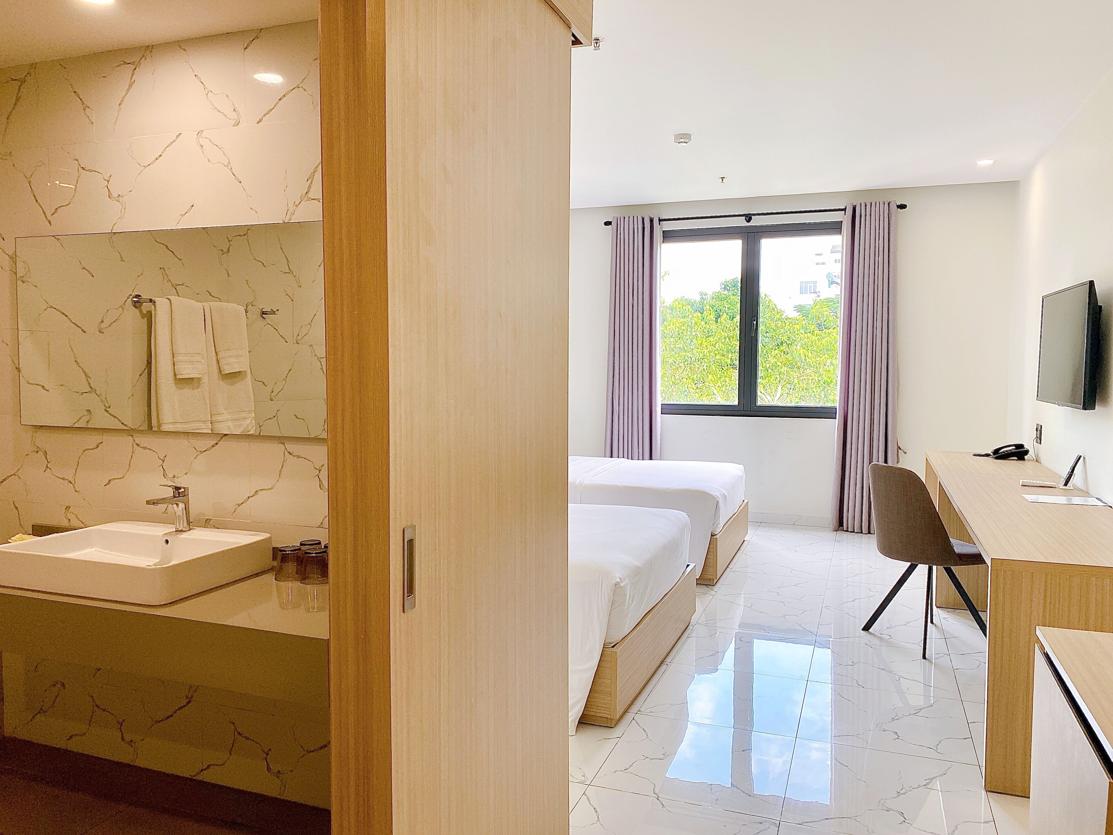 Quarto de hotel com banheiro de mármore, duas camas, uma escrivaninha e uma televisão.