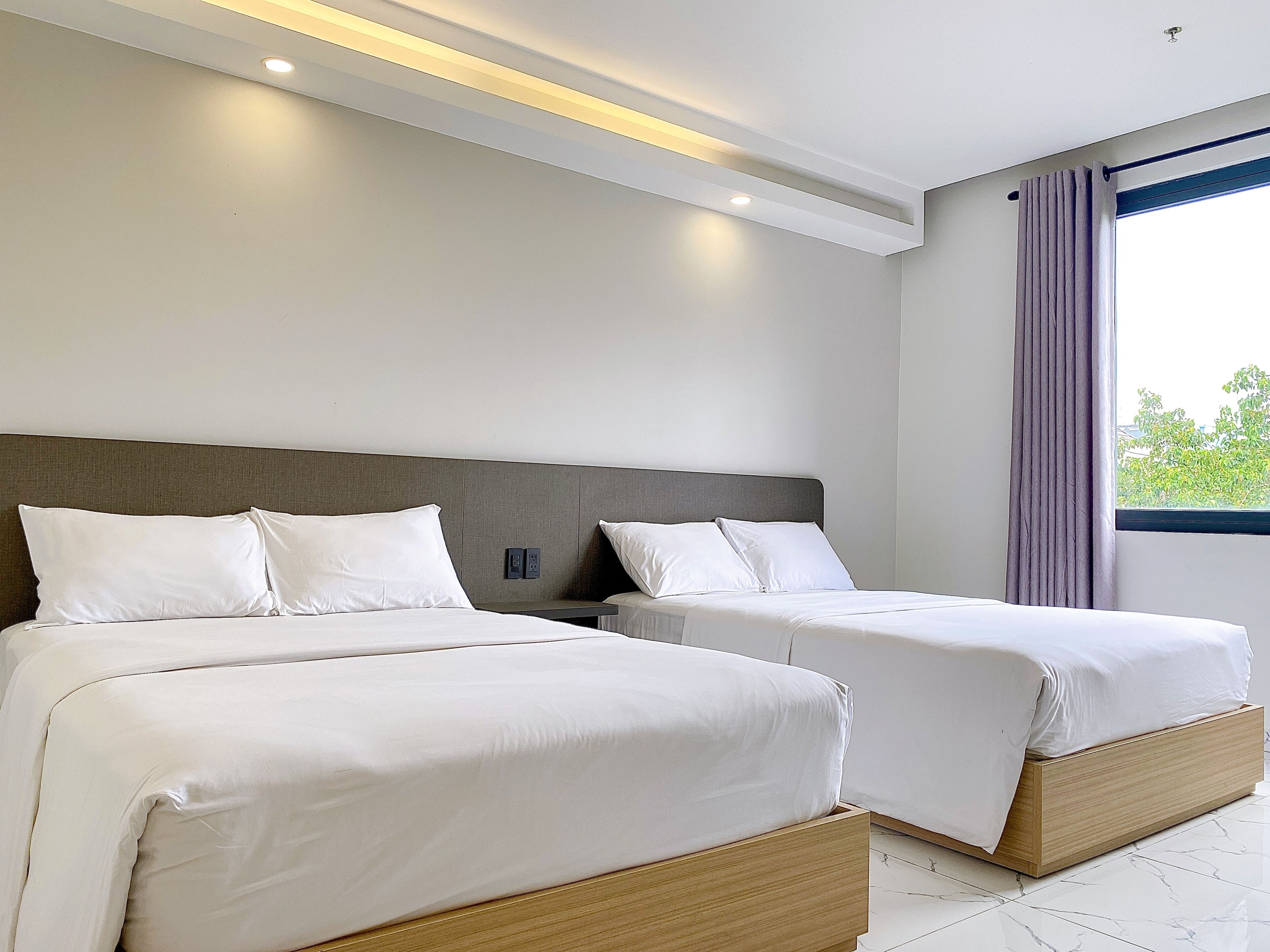 Quarto de hotel moderno com duas camas, uma janela grande com cortinas e iluminação embutida no teto.