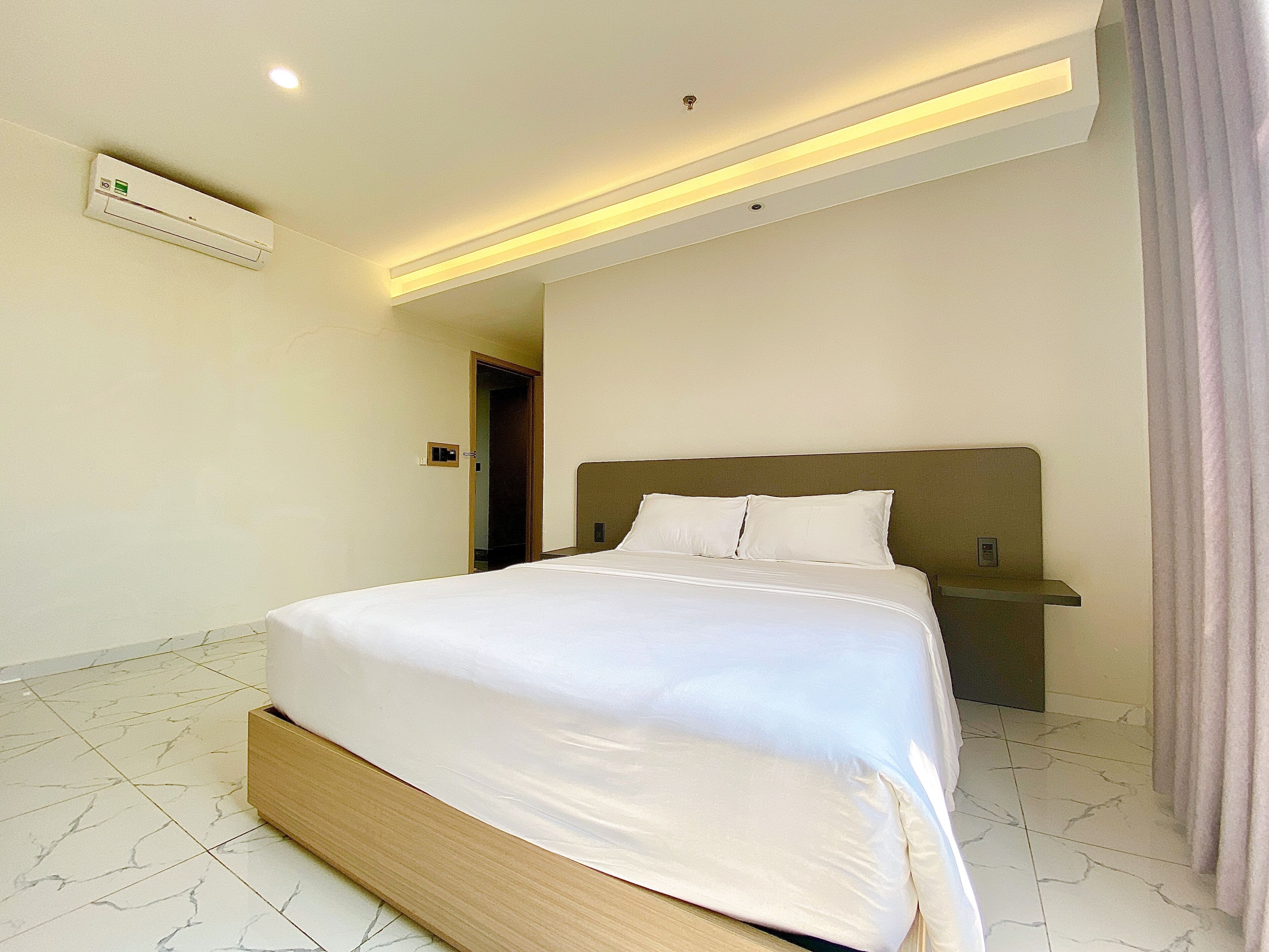 A hotel room with a large bed, a nightstand, a wall-mounted air conditioner, and a door leading to another room.