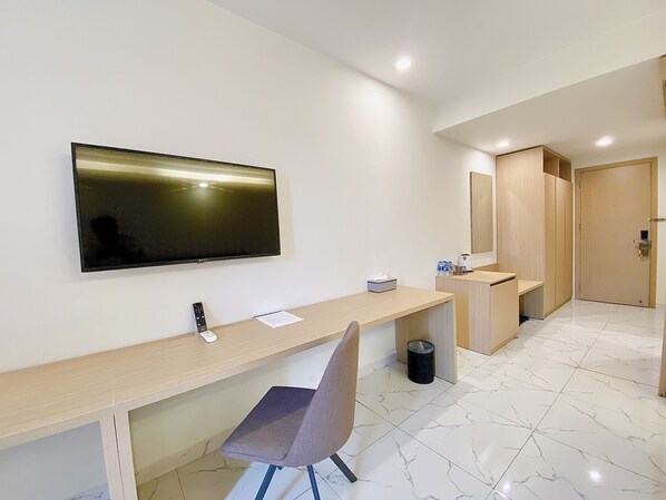 Una habitación de hotel moderna con un televisor de pantalla plana montado en la pared, un escritorio de madera con una silla, un cesto de basura pequeño y un armario de madera.