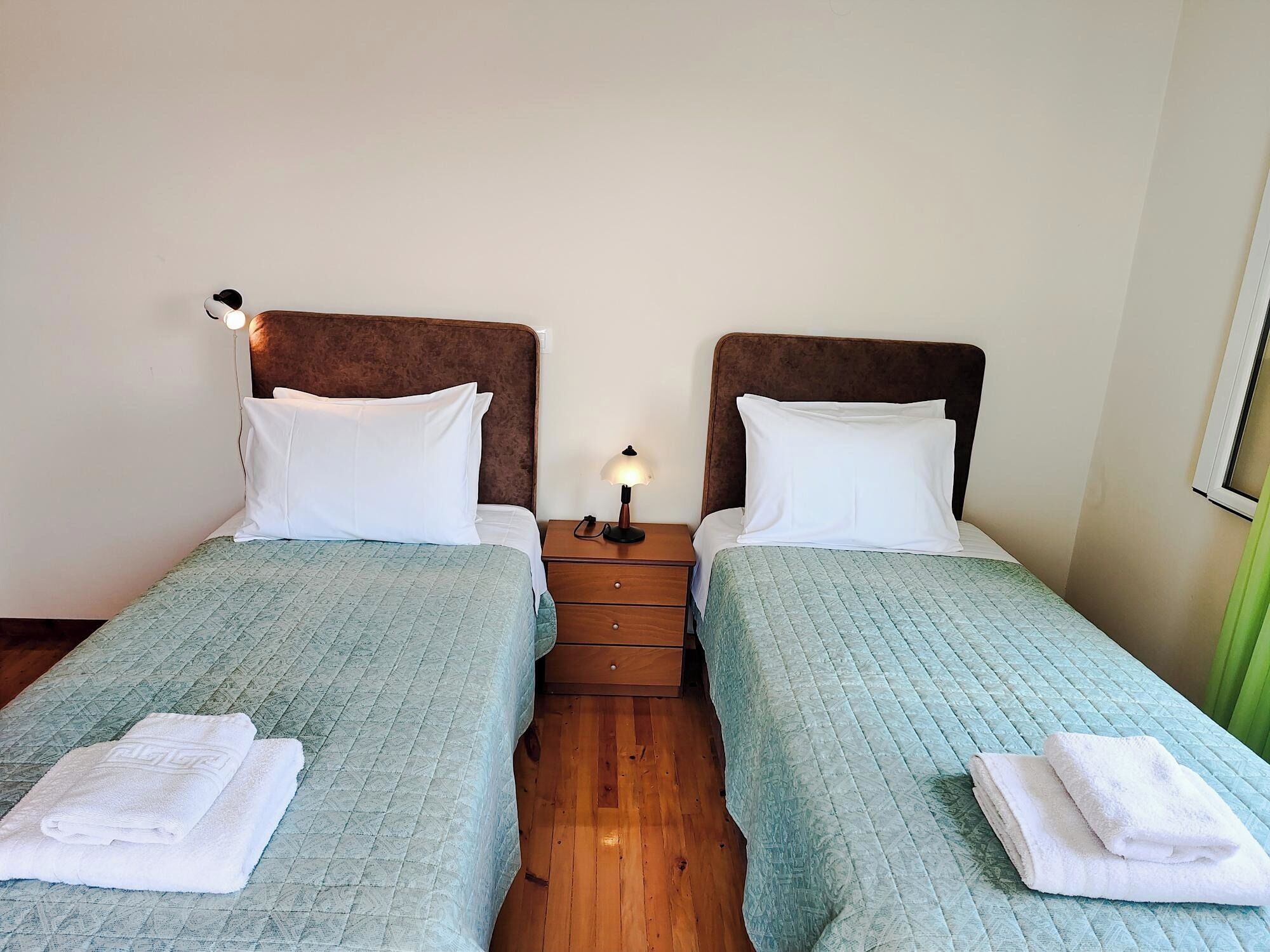 A double room with two beds, each with a headboard, a bedside table with a lamp, and a folded towel on each bed.