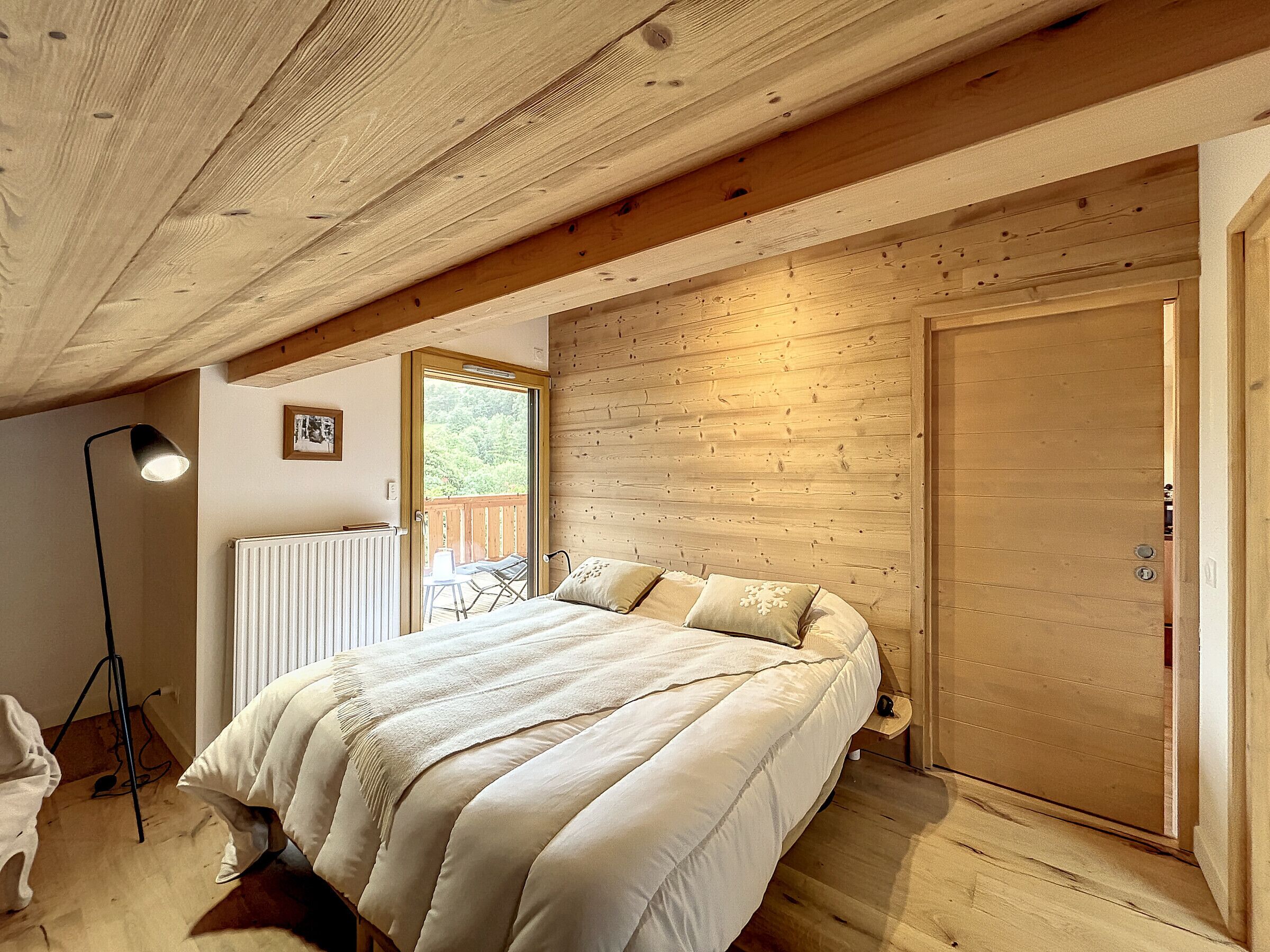 A bedroom with a wooden ceiling, a bed with white bedding, a radiator, and a door leading to an outdoor area.