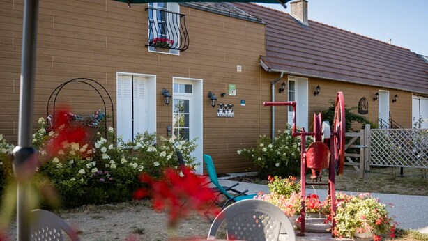 Un bâtiment à la façade marron, aux volets blancs et au toit rouge. On y trouve des chaises et une table d’extérieur, cette dernière étant recouverte d’une housse décorative.
