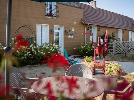 A building with a brown facade, white shutters, and a red roof. There are outdoor chairs and a table with a decorative cover.. Charming Riverside Gîte: Family-friendly, Horse-friendly, Perfect for Cyclists