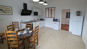Une cuisine avec des meubles blancs, une table à manger avec des chaises, un luminaire noir au plafond et un réfrigérateur.
