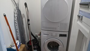 Une machine à laver et un sèche-linge superposés dans une buanderie, avec un balai et une serpillière appuyés contre le mur.