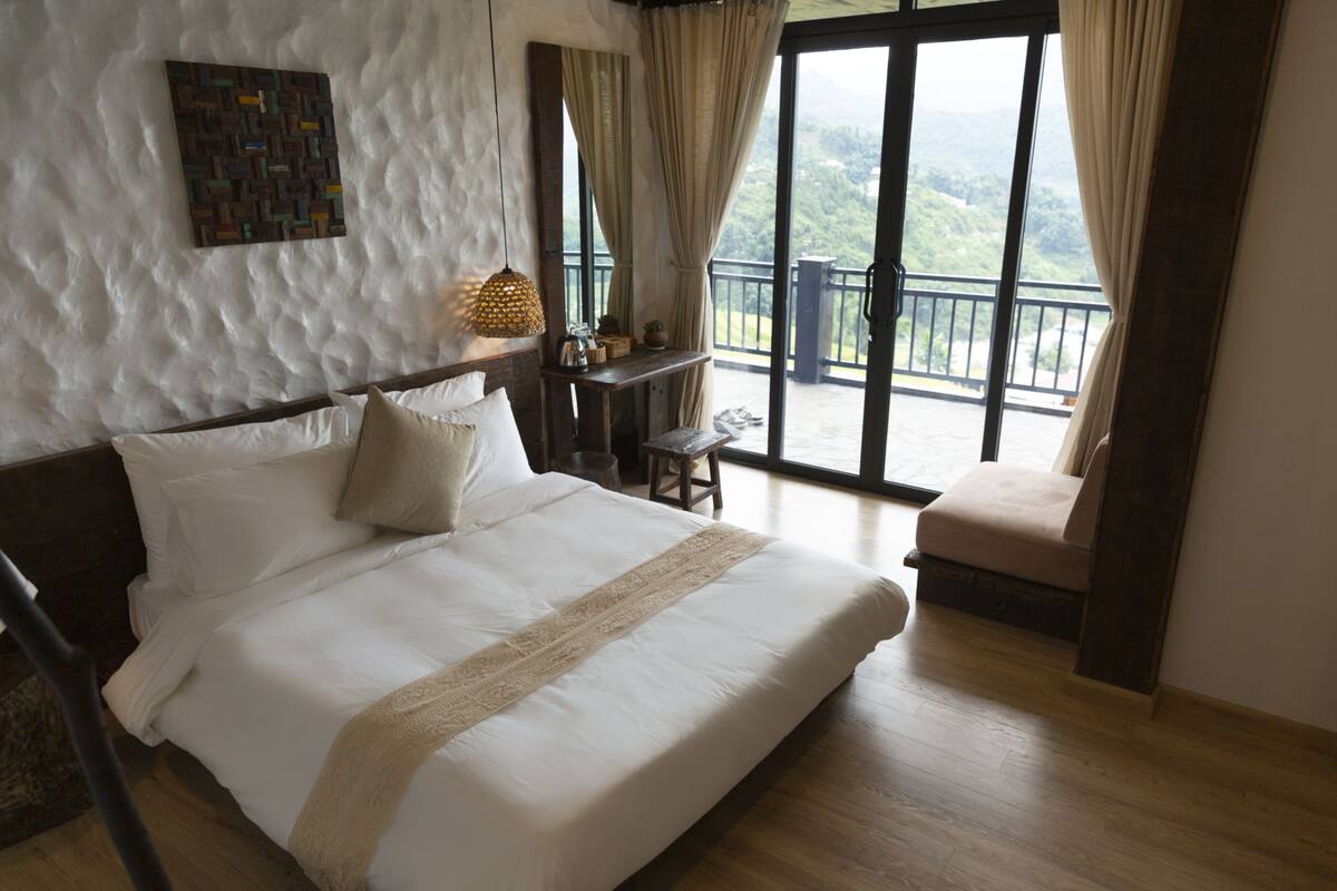A bedroom with a large bed, a wooden nightstand, a small table, and a view of a balcony with a railing.