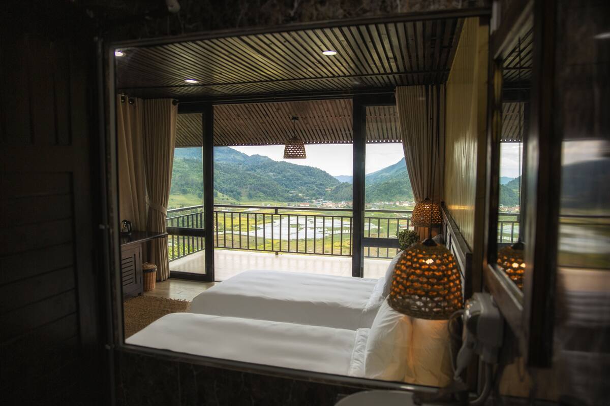 A bedroom with a large bed, a view of a mountainous landscape, and a balcony with a railing.