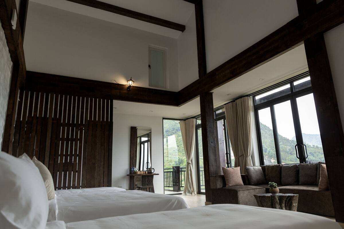 A bedroom with two beds, a wooden headboard, a balcony with a view, and a seating area with a table.
