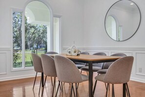 A dining area with a wooden table, six chairs, a round mirror, and a view of a garden through large windows.