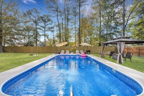A rectangular swimming pool with a wooden deck surrounding it, a covered seating area with chairs, and a wooden fence in the background.