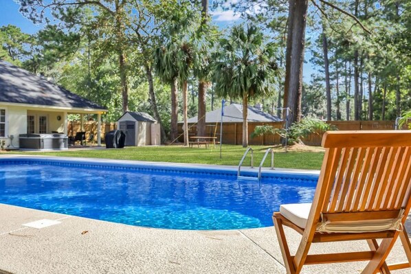 A swimming pool with a wooden chair, surrounded by trees and a house.
