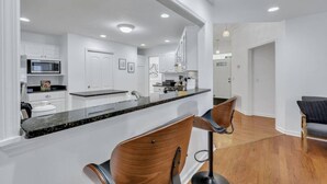 A modern kitchen with white cabinets, a granite countertop, and a bar area with two wooden bar stools. - Savannah 5BR Pool, Jacuzzi, Firepit, Sports, Games (Savannah)