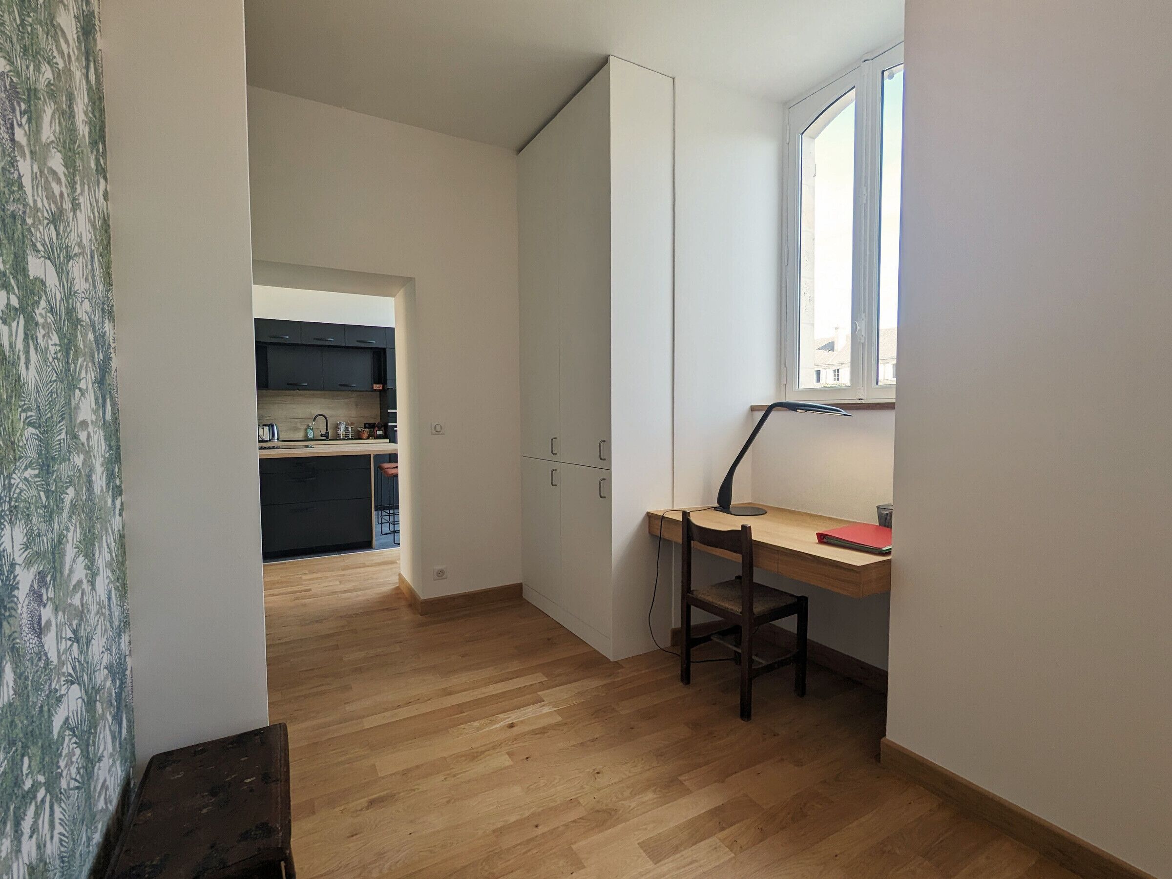 Een compacte kamer met een houten bureau, een stoel en een raam. In de achtergrond is een keuken te zien.