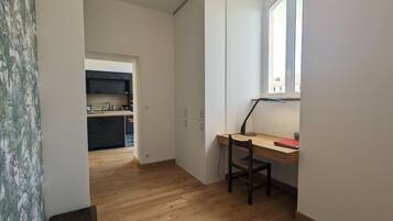 Een compacte kamer met een houten bureau, een stoel en een raam. In de achtergrond is een keuken te zien.