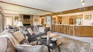 A spacious living room with a large sofa, a coffee table, and a fireplace. There is a kitchen area with wooden cabinets and a bar counter. - 3BR Condo Retreat w/ Seasonal Pool & Hot Tub, Balcony, Views & Near Ober Mtn (Gatlinburg)
