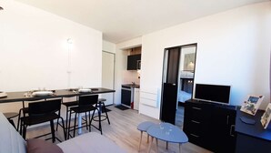 Een ruimtelijk ingerichte leefruimte met kitchenette, eethoek, bank en televisie.