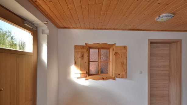 A room with a wooden ceiling, a window with wooden shutters, a wooden door, and a tiled floor. - Holzhammer Hof (Bayerbach)