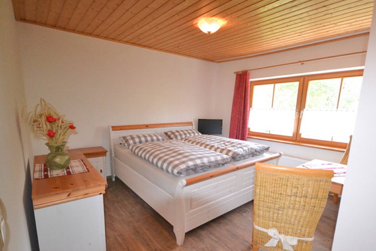 A bedroom with a bed, a chair, a small table, a window, and a wall-mounted TV.