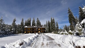 A snow-covered landscape with a building featuring a symmetrical facade and multiple balconies, surrounded by tall evergreen trees.