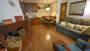 A cozy living room with a sofa, a dining table, and chairs. There is a wooden floor and a kitchen area in the background.
