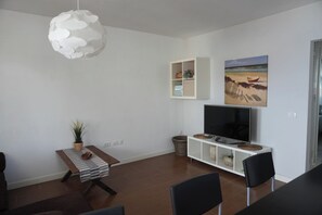 Ein modernes Wohnzimmer mit Flachbildfernseher, einer Essecke mit Stühlen, einer weißen Kommode und einem Strandgemälde.
