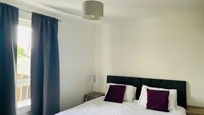 Een net opgemaakt bed met witte en paarse kussens, een bedlampje en een raam met blauwe gordijnen.