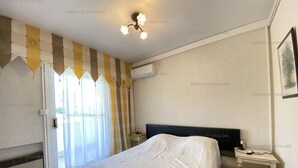 A bedroom with a bed covered in white, a nightstand, a wall-mounted air conditioner, and a window with curtains.