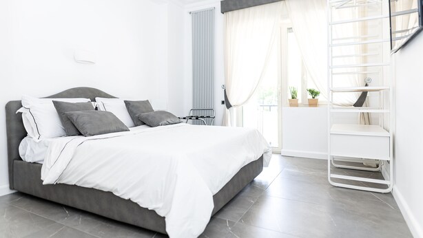 A modern bedroom with a grey sofa bed, white bedding, a white metal shelf unit, and a window with sheer curtains. - Enjoy your holiday in Naples in this brand new apartment (Napoli)