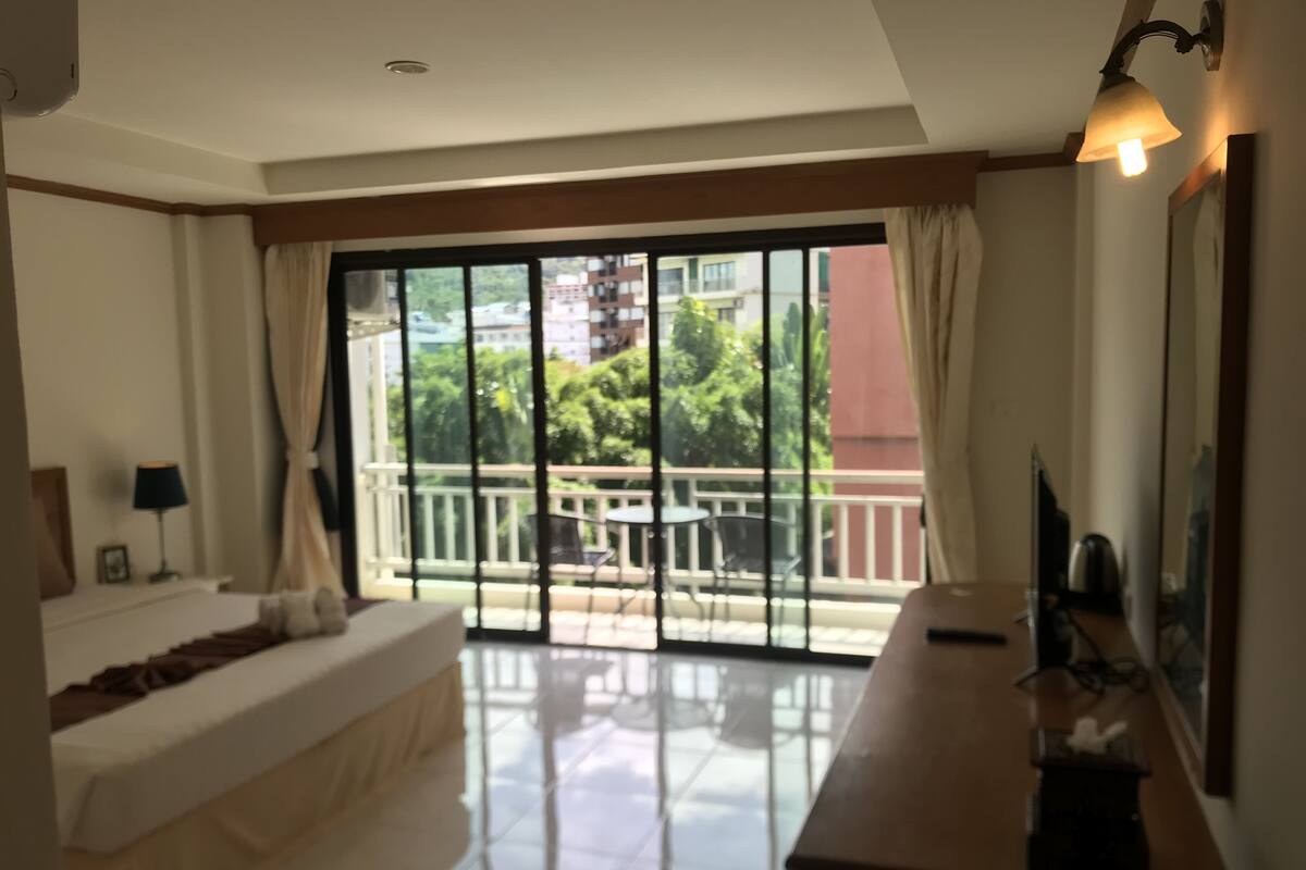 A bedroom with a large sliding glass door leading to a balcony, a bed, a bedside table with a lamp, and a TV on a stand.