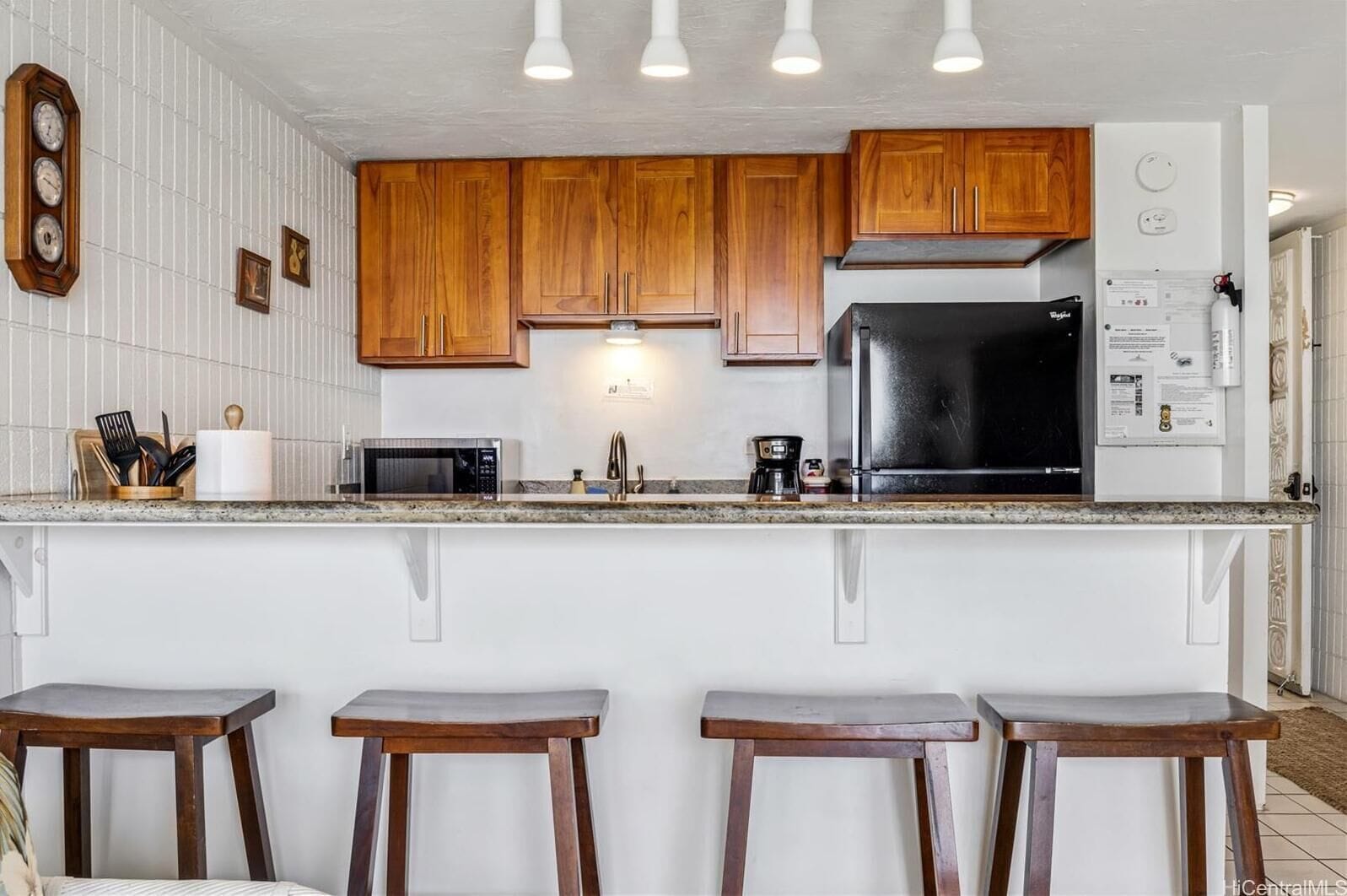 A kitchen with wooden cabinets, a black refrigerator, a microwave, and a coffee maker. There are three wooden stools at the counter.
